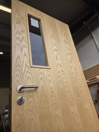 Ash veneer fire door featuring a vertical rectangular glazed vision panel, a satin stainless steel lever handle, and a thumb-turn lock.